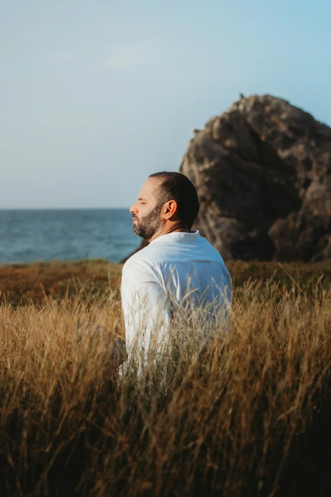 A man sits peacefully in the grass, gazing at the ocean waves under a clear blue sky.