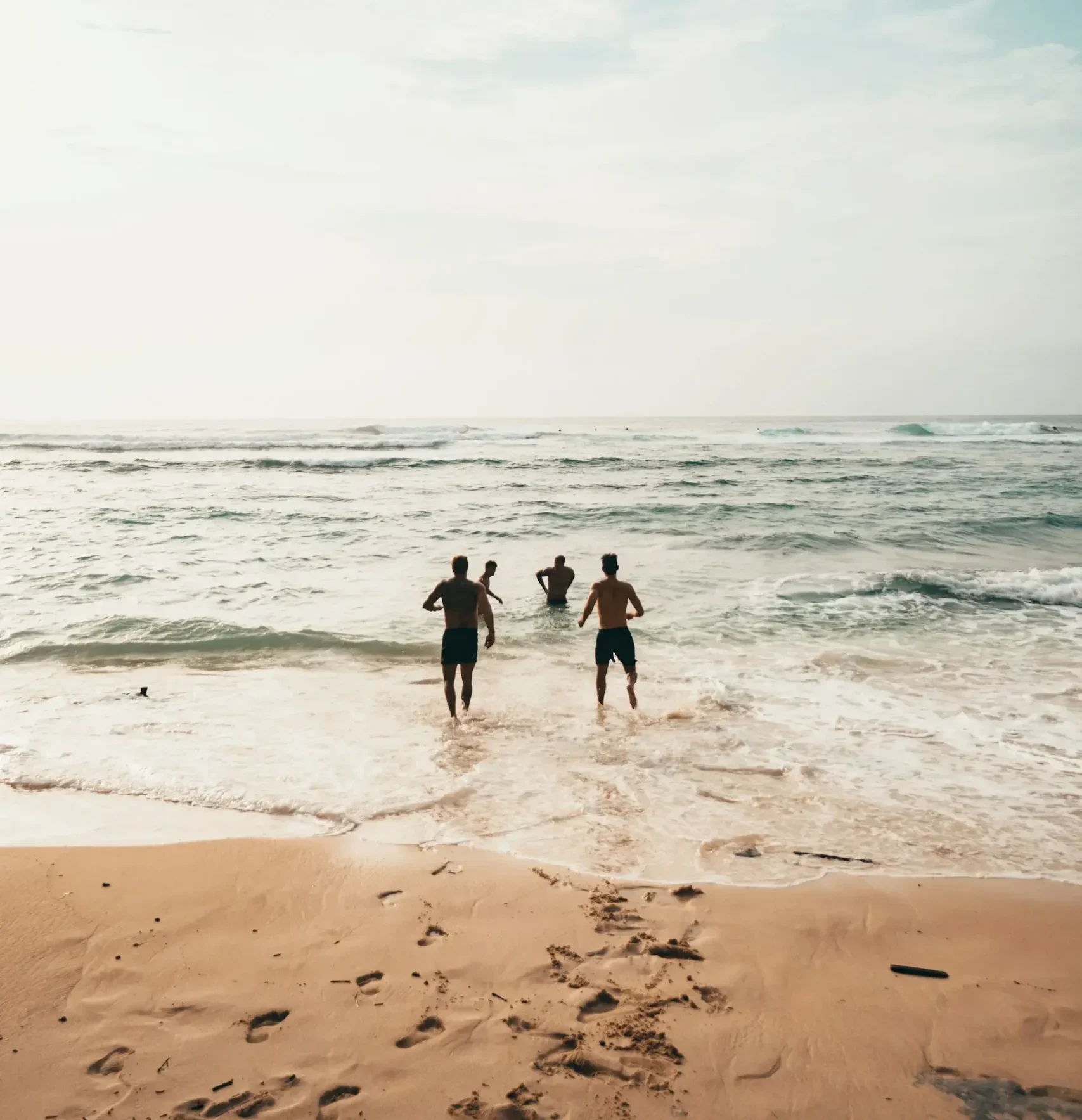 Three people joyfully running into the ocean waves at a sunny beach, splashing water around them.