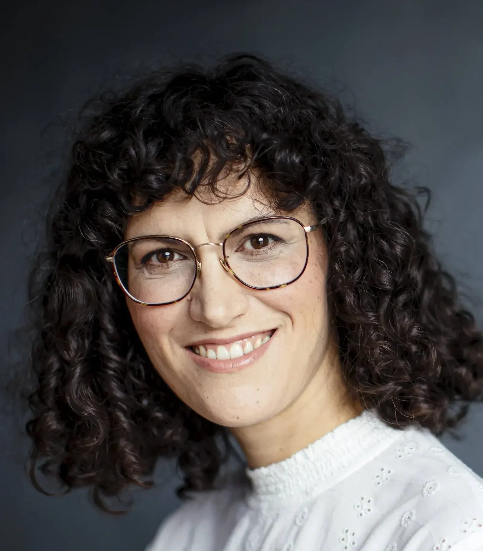 A woman with curly hair and glasses smiles warmly at the camera.
