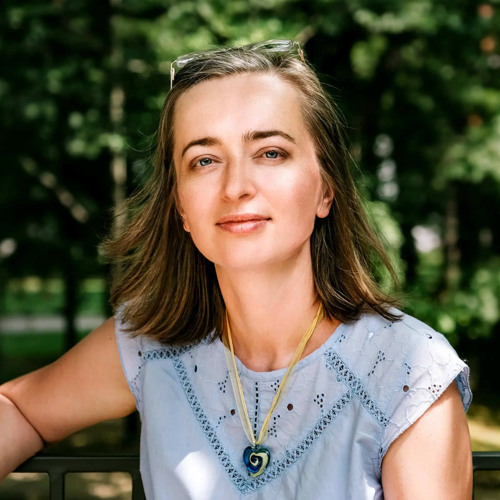 A woman in a blue top and necklace smiles warmly at the camera, showcasing her stylish outfit.