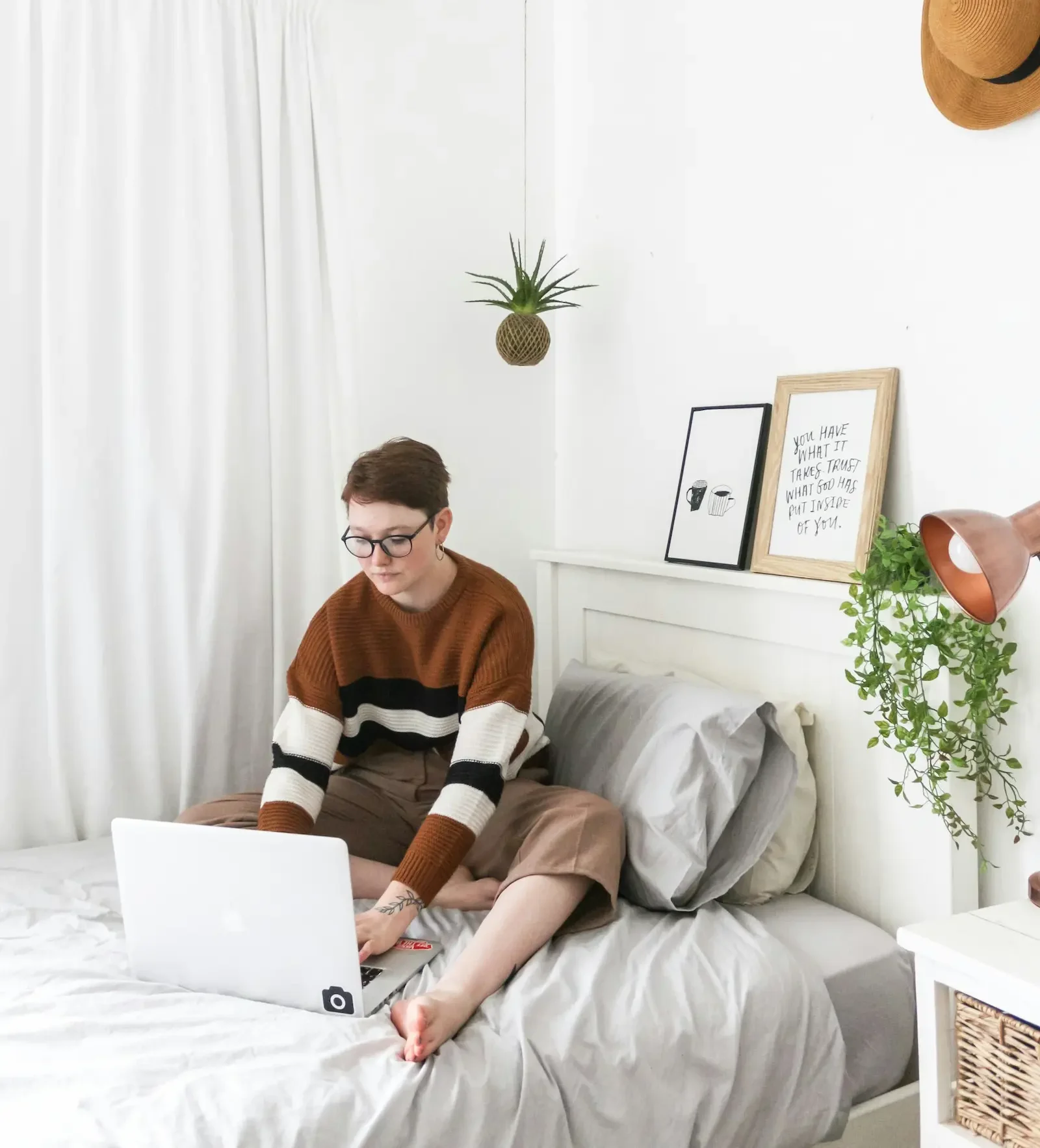 A woman sitting on a bed, focused on her laptop, surrounded by soft pillows and a cozy atmosphere.
